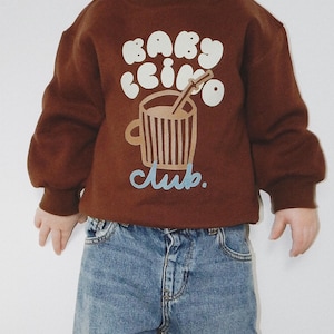 May include: A brown sweatshirt with the text "BABY CINO Club" in white and light blue. The graphic features a coffee cup with a straw. The sweatshirt is paired with blue denim jeans.