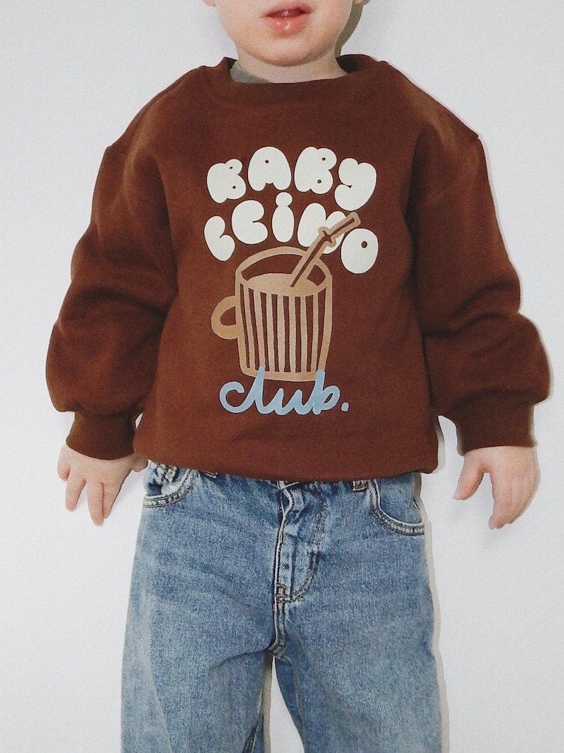 May include: A brown sweatshirt with the text "BABY CINO Club" in white and light blue. The graphic features a coffee cup with a straw. The sweatshirt is paired with blue denim jeans.