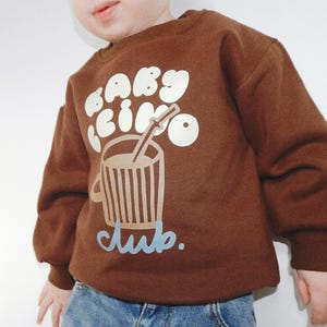 May include: Brown sweatshirt with the text "baby cino club" in white and light blue. The graphic features a coffee cup with a straw. The sweatshirt is worn by a child.