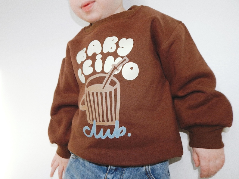 May include: Brown sweatshirt with the text "baby cino club" in white and light blue. The graphic features a coffee cup with a straw. The sweatshirt is worn by a child.