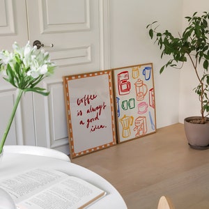 May include: Two framed prints on a white wall. The print on the left has a white background with orange and brown checkered border. The text on the print reads "Coffee is always a good idea." The print on the right has a white background with a brown border. The print features colorful illustrations of coffee makers, mugs, and coffee beans.