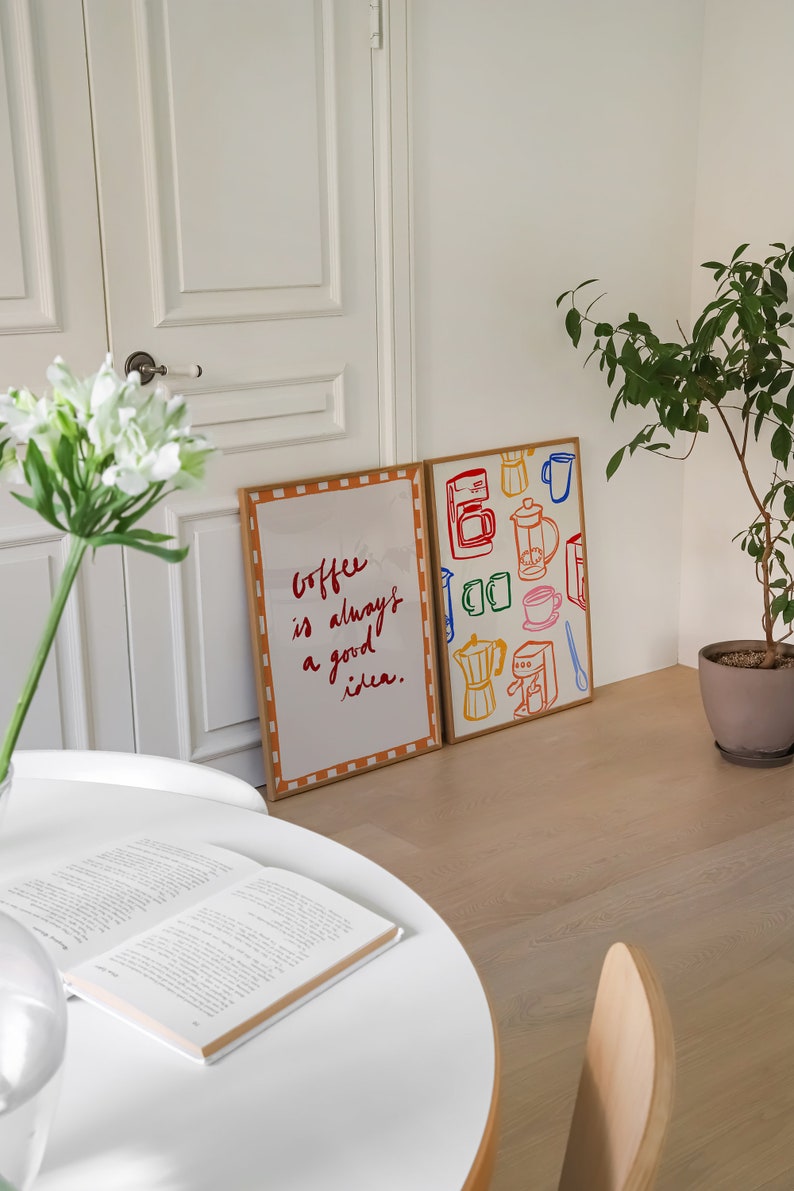 May include: Two framed prints on a white wall. The print on the left has a white background with orange and brown checkered border. The text on the print reads "Coffee is always a good idea." The print on the right has a white background with a brown border. The print features colorful illustrations of coffee makers, mugs, and coffee beans.
