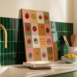 May include: A framed art print featuring a grid of colorful tomato illustrations. The artwork includes the word "POMODORO" and is set against a backdrop of pink, beige, and brown squares. The frame is made of light-colored wood.