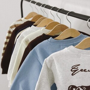 May include: A selection of children's sweatshirts in various colors, hanging on wooden hangers. The sweatshirts include a light blue, gray, and brown one. The gray sweatshirt has a rocking horse graphic and the word "Gee" printed on it.