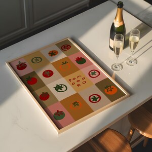 May include: A framed artwork featuring a grid of colorful squares with tomato illustrations and the word "POMODORO". A bottle of champagne and two filled glasses sit nearby. The artwork is on a white countertop.