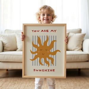 May include: A framed art print featuring a smiling sun illustration in shades of orange and yellow, with the text "YOU ARE MY SUNSHINE" in red. The background has vertical gray stripes. The frame is a light wood color.