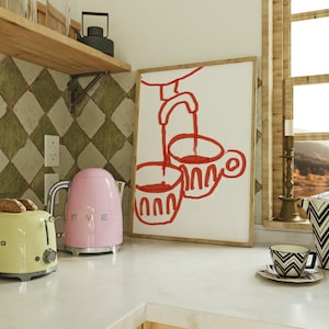 May include: A pink electric kettle and a yellow toaster on a white countertop. A framed illustration of two red cups with a red faucet pouring liquid into the cups hangs on the wall behind the appliances.