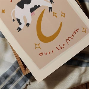 May include: A whimsical art print featuring a cow floating over a golden crescent moon. The cow is black and white with pink udders. The words "over the moon" are written in red script. Gold stars are scattered around the moon.