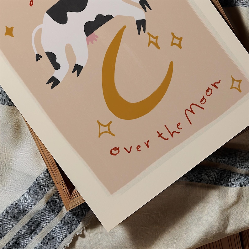 May include: A whimsical art print featuring a cow floating over a golden crescent moon. The cow is black and white with pink udders. The words "over the moon" are written in red script. Gold stars are scattered around the moon.