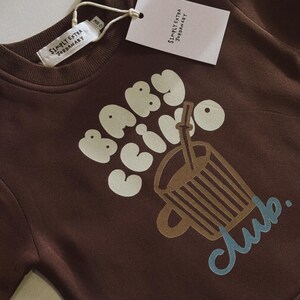 May include: A brown sweatshirt with the words "BABY COFFEE CLUB" in white and a graphic of a coffee cup. The sweatshirt has a tag that reads "SIMPLY EXTRA JORDANARY".