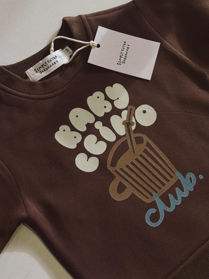 May include: A brown sweatshirt with the words "BABY COFFEE CLUB" in white and a graphic of a coffee cup. The sweatshirt has a tag that reads "SIMPLY EXTRA JORDANARY".