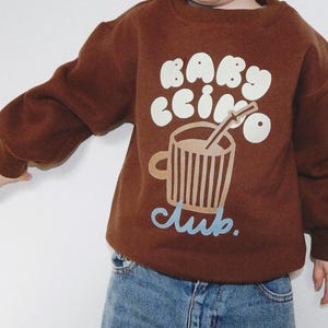 May include: A brown sweatshirt with the words "Babyccino Club" in white and light blue. The graphic features a coffee cup with a straw. The sweatshirt has long sleeves and is worn with blue jeans.