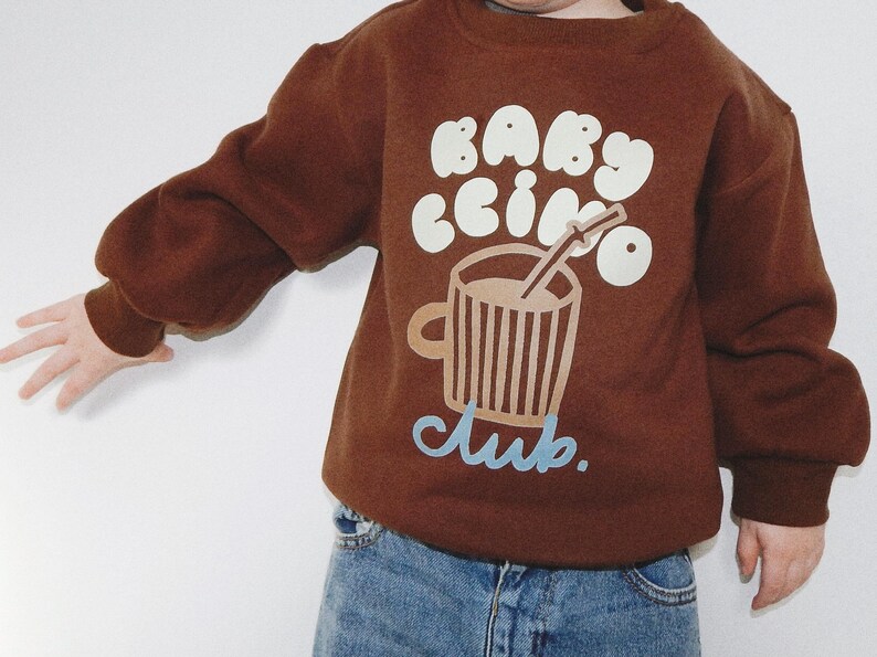 May include: A brown sweatshirt with the words "Babyccino Club" in white and light blue. The graphic features a coffee cup with a straw. The sweatshirt has long sleeves and is worn with blue jeans.