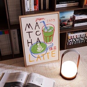 Peut inclure: Une illustration encadr&eacute;e d'un latte au matcha avec le texte "Japan 1999" et "抹茶 LATTE" en japonais et en anglais. L'illustration est dans un style cartoon color&eacute; avec une tasse verte de latte au matcha et un verre de matcha glac&eacute;.