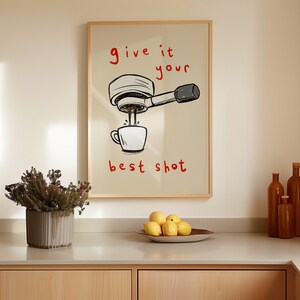 以下が含まれることがあります： コーヒーをテーマにしたイラストが描かれた額入りのプリント。アートワークは、カップに注がれるコーヒーマシンを描いており、赤い文字で「give it your best shot」と書かれています。プリントは、カウンタートップの上にある明るい色の壁に表示されています。