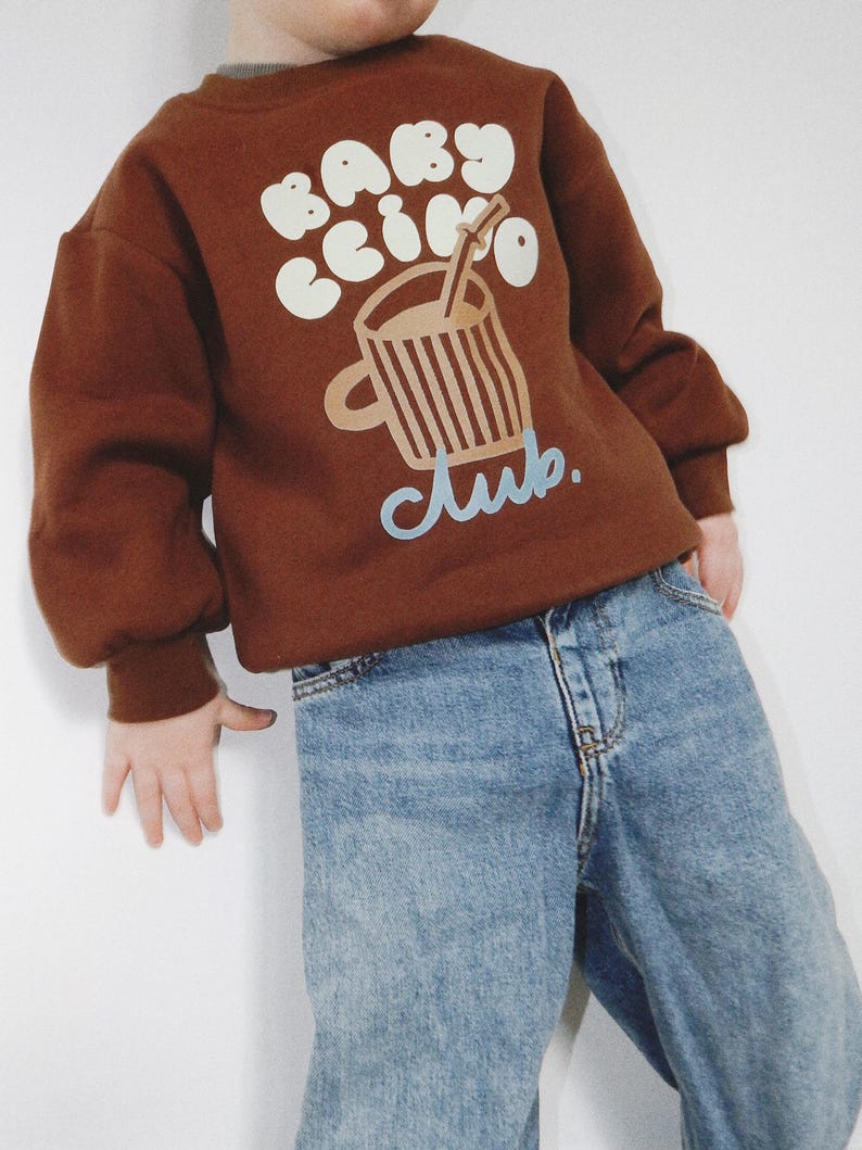 May include: A brown sweatshirt with the text "BABY CCINO CLUB" in white and light blue. The sweatshirt features a graphic of a coffee cup with a straw. The child is wearing light blue denim jeans.