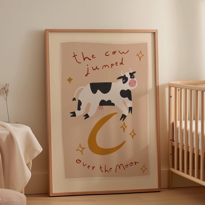 May include: Framed art print featuring a whimsical illustration of a cow jumping over a crescent moon. The cow is black and white with a pink nose, and the text reads "the cow jumped over the moon". The background is a soft beige color.