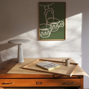 May include: A wooden drawer cabinet with five drawers, a white marble lamp, a book titled "Original Bauhaus", and a green framed illustration of two cups with a spout pouring into them.