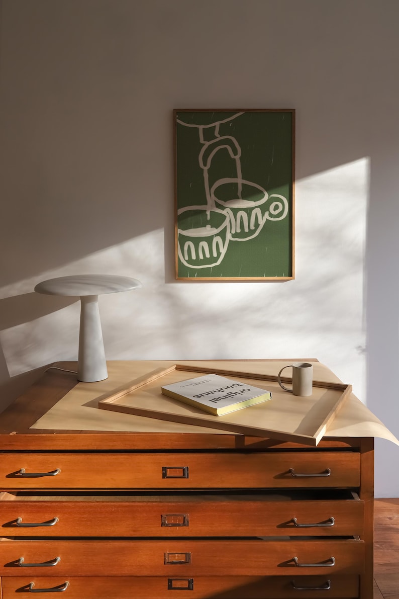 May include: A wooden drawer cabinet with five drawers, a white marble lamp, a book titled "Original Bauhaus", and a green framed illustration of two cups with a spout pouring into them.