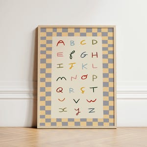 May include: A framed alphabet print featuring colorful handwritten letters from A to Z. The letters are in various colors, set against a cream background. The frame has a light wood finish, and the border is a checkered pattern in gray and yellow.