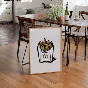 K&ouml;nnte beinhalten: Ein gerahmter Druck einer Schwarzwei&szlig;illustration einer Schachtel Pommes mit dem McDonald's-Logo. Die Pommes sind gelb und die Schachtel ist wei&szlig; mit schwarzem Umriss. Der Druck h&auml;ngt an einer Wand in einem Esszimmer.