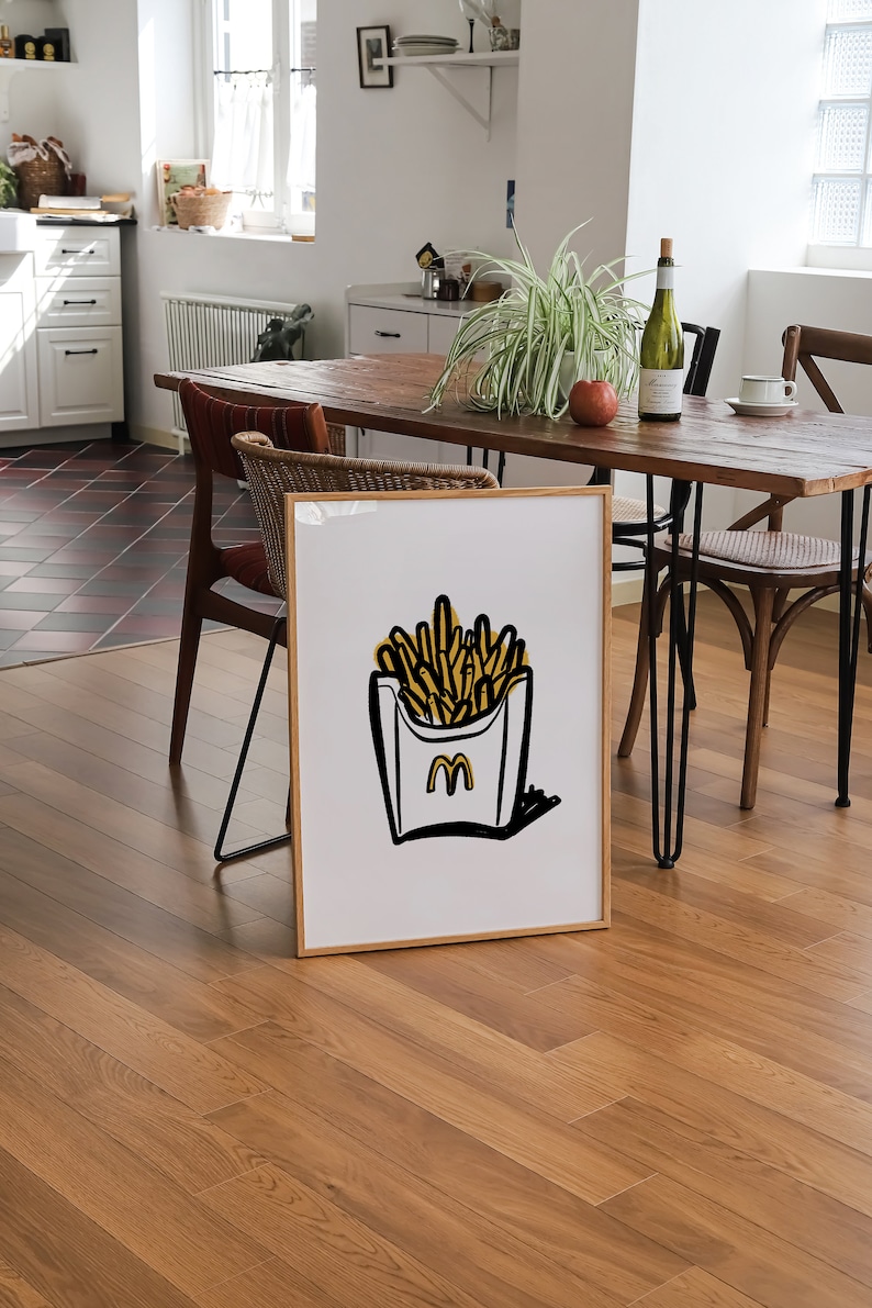 K&ouml;nnte beinhalten: Ein gerahmter Druck einer Schwarzwei&szlig;illustration einer Schachtel Pommes mit dem McDonald's-Logo. Die Pommes sind gelb und die Schachtel ist wei&szlig; mit schwarzem Umriss. Der Druck h&auml;ngt an einer Wand in einem Esszimmer.