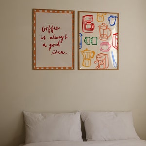 May include: Two framed prints on a white wall above a white bed with two white pillows. The print on the left says "Coffee is always a good idea." The print on the right has a colorful pattern of coffee makers, mugs, and coffee beans.