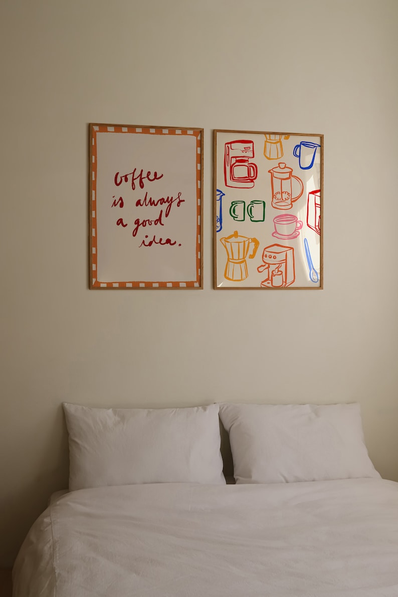 May include: Two framed prints on a white wall above a white bed with two white pillows. The print on the left says "Coffee is always a good idea." The print on the right has a colorful pattern of coffee makers, mugs, and coffee beans.