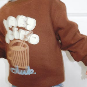 May include: A brown sweatshirt with a graphic of a drink and the words "baby drink club." The text and drink graphic are in white and light brown. The sweatshirt is worn with blue jeans.