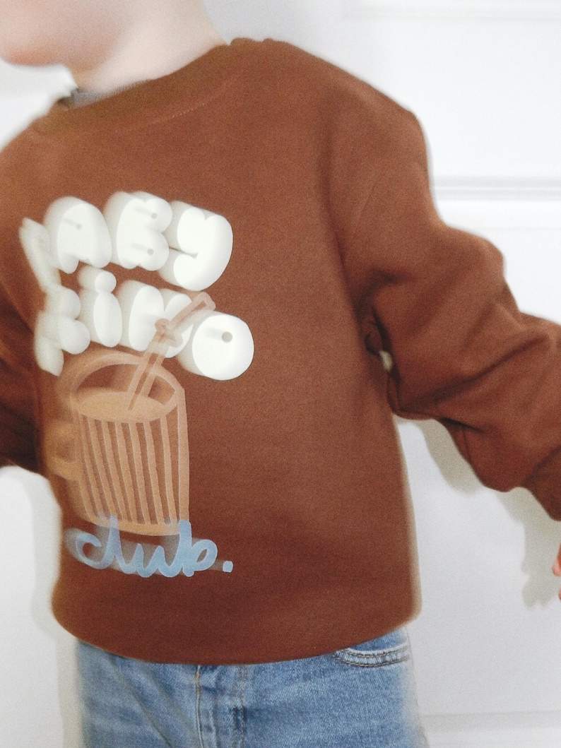 May include: A brown sweatshirt with a graphic of a drink and the words "baby drink club." The text and drink graphic are in white and light brown. The sweatshirt is worn with blue jeans.
