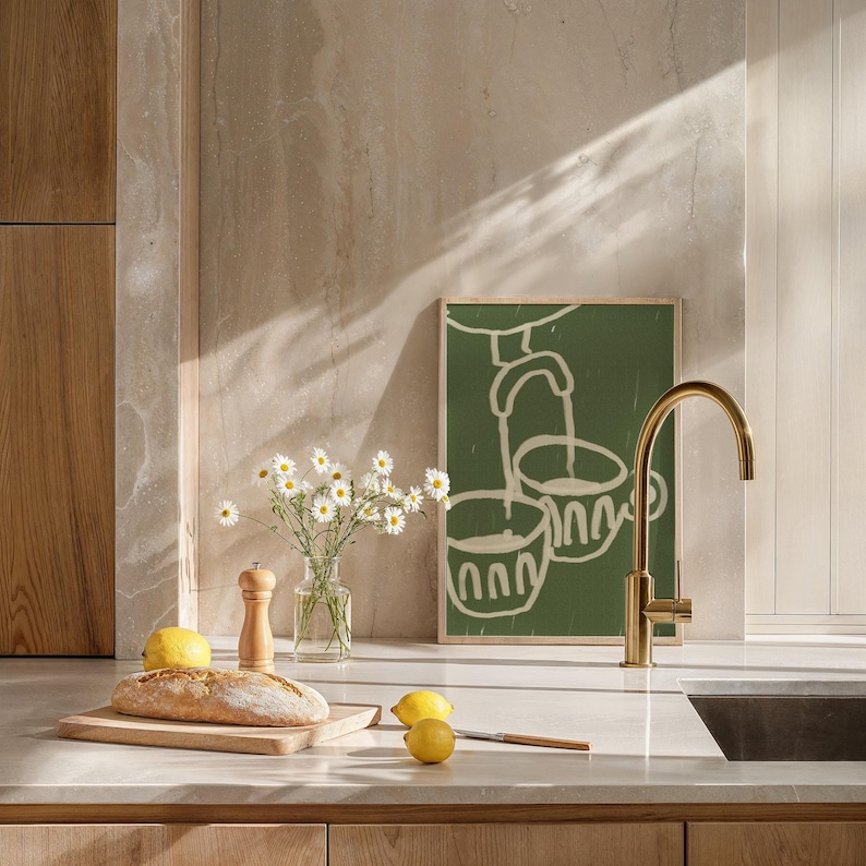May include: A kitchen countertop with a wooden cutting board, a loaf of bread, lemons, a salt and pepper shaker, a bouquet of white daisies in a glass vase, and a green framed print of a stylized illustration of a faucet pouring water into two cups. A gold faucet is mounted to the countertop.