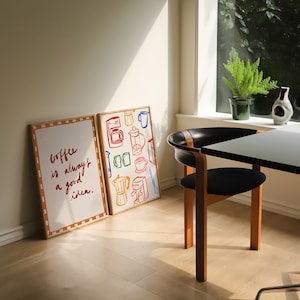 May include: Two framed prints on a light wood floor. The print on the left says "Coffee is always a good idea." The print on the right features a colorful illustration of coffee brewing equipment.