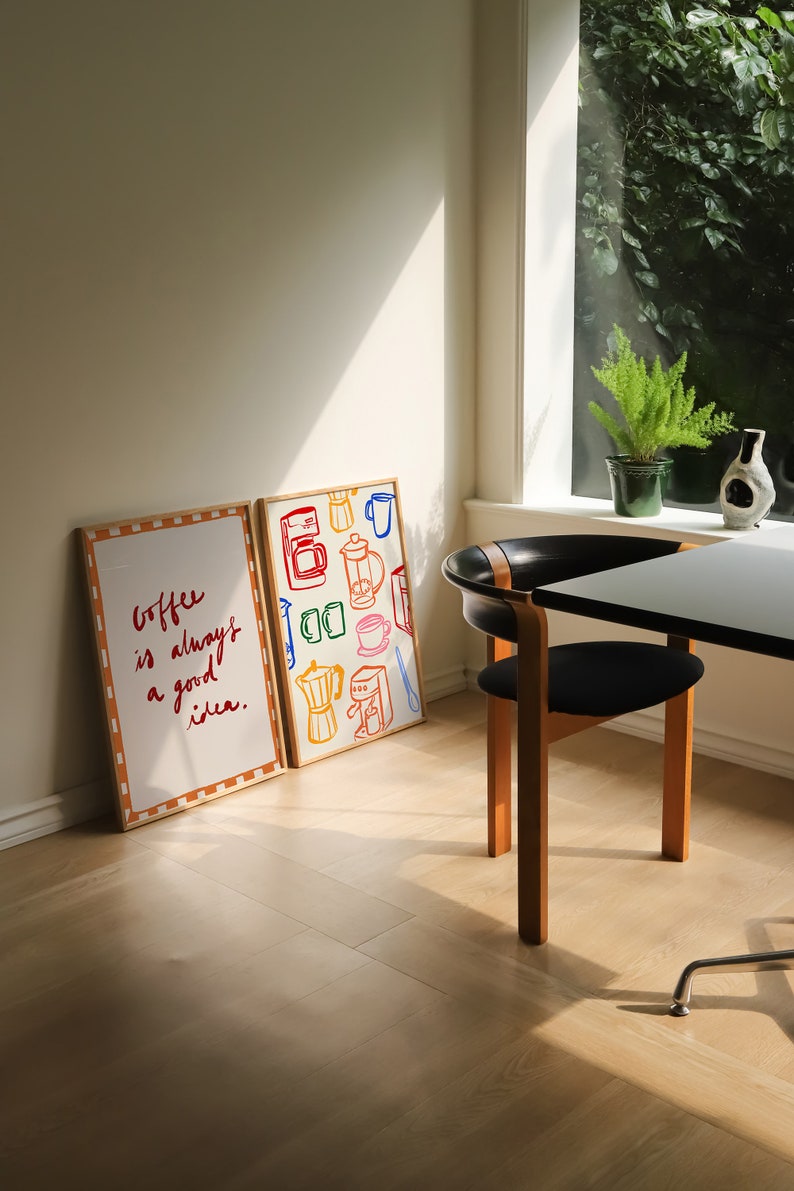 May include: Two framed prints on a light wood floor. The print on the left says "Coffee is always a good idea." The print on the right features a colorful illustration of coffee brewing equipment.