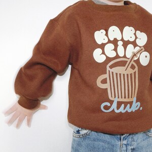 May include: A brown sweatshirt with the words "BABY CCINO" in white bubble letters and a graphic of a coffee cup with a straw. The word "club" is written in light blue script below the cup.