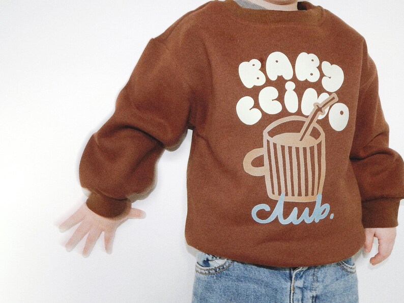 May include: A brown sweatshirt with the words "BABY CCINO" in white bubble letters and a graphic of a coffee cup with a straw. The word "club" is written in light blue script below the cup.