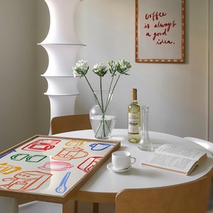 May include: A white table with a wooden frame and a white tabletop. On the table is a framed print with the text "Coffee is always a good idea." There is also a book, a white coffee cup, a bottle of wine, a vase with white flowers, and a tray with a colorful illustration of coffee makers and cups.