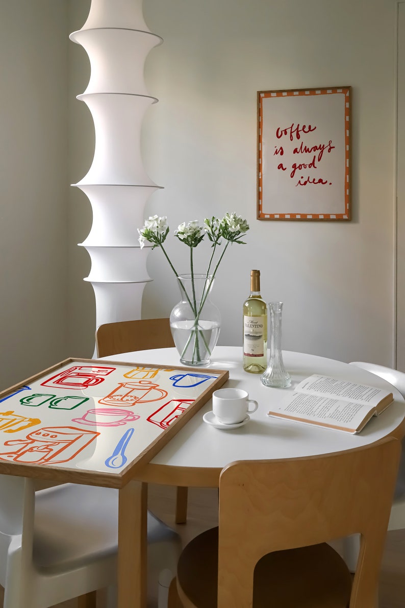 May include: A white table with a wooden frame and a white tabletop. On the table is a framed print with the text "Coffee is always a good idea." There is also a book, a white coffee cup, a bottle of wine, a vase with white flowers, and a tray with a colorful illustration of coffee makers and cups.
