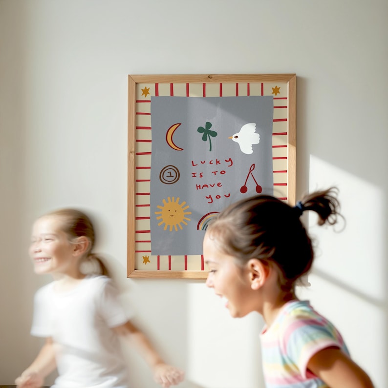 May include: A framed print with a gray background and red and white stripes around the border. The print features a variety of hand-drawn illustrations, including a crescent moon, a four-leaf clover, a bird, a sun, a rainbow, and a cherry. The text on the print reads "Lucky is to have you".