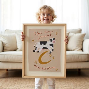May include: A framed art print featuring a whimsical illustration of a cow jumping over a crescent moon. The cow is black and white, with the text "the cow jumped over the moon" in a handwritten style. The print has a beige background and gold accents.