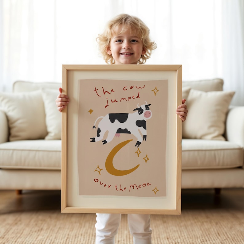 May include: A framed art print featuring a whimsical illustration of a cow jumping over a crescent moon. The cow is black and white, with the text "the cow jumped over the moon" in a handwritten style. The print has a beige background and gold accents.