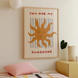 May include: A framed art print featuring a smiling sun illustration in shades of orange and brown. The words "YOU ARE MY SUNSHINE" are printed in red. The background has vertical gray stripes on a cream color.