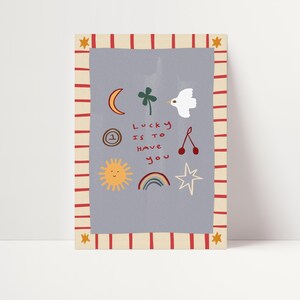 May include: A light blue card with red and white stripes around the border. The card has a variety of illustrations including a crescent moon, a four-leaf clover, a bird, a cherry, a sun, a rainbow, and a star. The text on the card reads "Lucky is to have you".