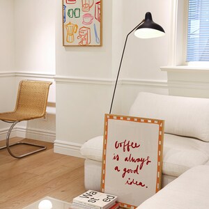 May include: A white couch with a throw pillow and a framed sign that reads "Coffee is always a good idea."  A black floor lamp with a white shade is next to the couch. A small table with a white lamp and a stack of books is in the foreground.