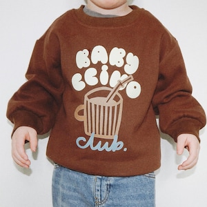 May include: A brown sweatshirt with the words "BABY ccino club" in white and light blue. The graphic features a cup of coffee with a straw. The child is wearing blue jeans.
