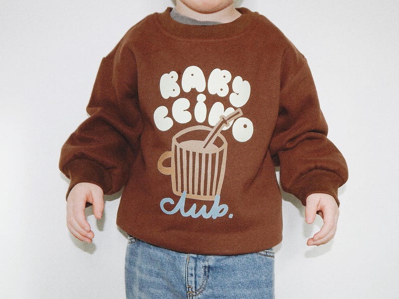 May include: A brown sweatshirt with the words "BABY ccino club" in white and light blue. The graphic features a cup of coffee with a straw. The child is wearing blue jeans.