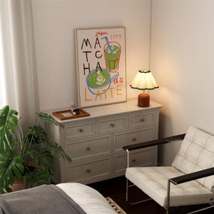 Peut inclure: Une commode blanche avec six tiroirs et des boutons dor&eacute;s. Une chaise blanche avec un cadre en m&eacute;tal noir est plac&eacute;e devant la commode. Une impression encadr&eacute;e avec le texte "Japan 1999 Matcha Latte" est accroch&eacute;e au-dessus de la commode. Une petite lampe de table avec un abat-jour blanc est pos&eacute;e sur la commode.