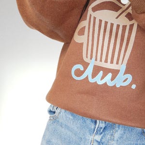 May include: Brown sweatshirt with a graphic of a coffee cup and the word "club" in light blue. The sweatshirt is paired with light blue denim jeans. The image is a close-up of the clothing.