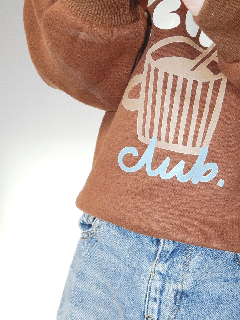 May include: Brown sweatshirt with a graphic of a coffee cup and the word "club" in light blue. The sweatshirt is paired with light blue denim jeans. The image is a close-up of the clothing.