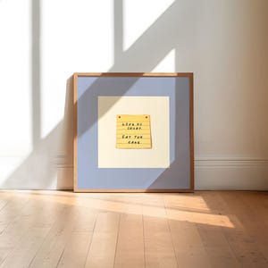 Pode incluir: Uma impressão artística emoldurada com um design de bloco de notas amarelo, exibindo o texto "LIFE IS SHORT. EAT THE CAKE." A obra de arte está sobre um fundo azul claro e creme, dentro de uma moldura de madeira. A impressão é exibida em um piso de madeira.
