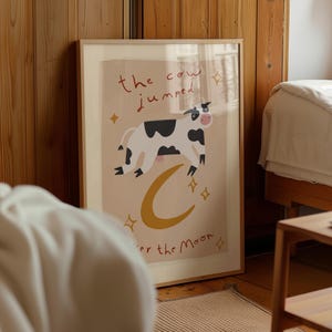 May include: Framed art print featuring a whimsical illustration of a cow jumping over a yellow crescent moon. The text "the cow jumped over the moon" is written in red and black. The artwork has a light beige background and is surrounded by a wooden frame.